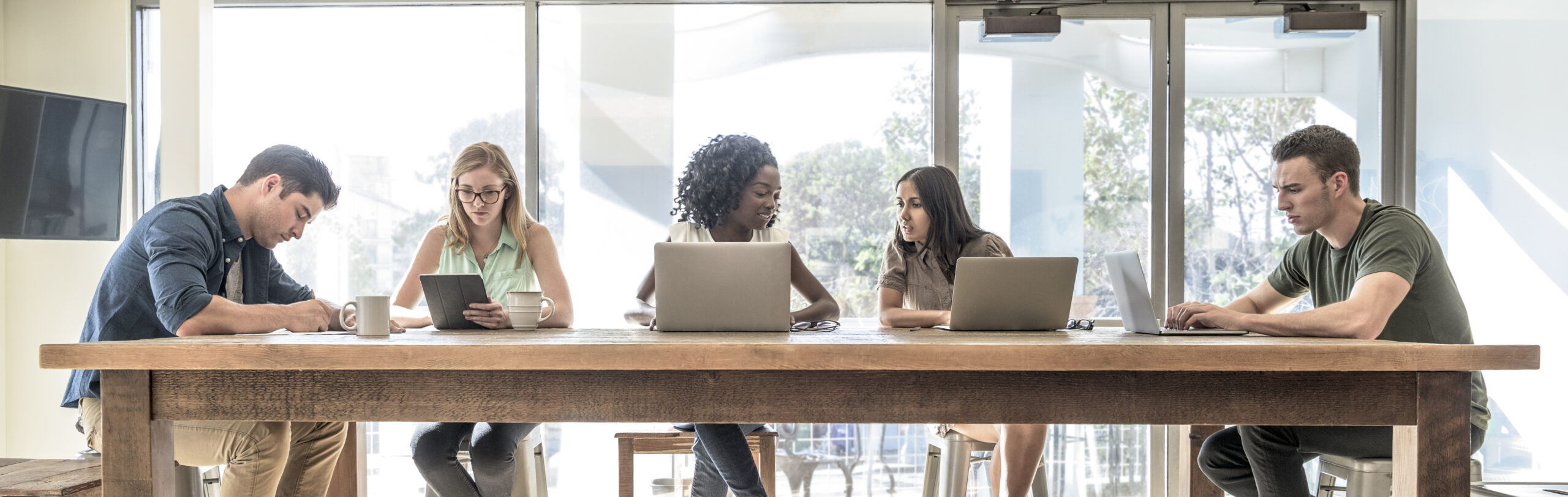 Panoramic composition of young people are sitting around a large table working together and individually in open plan office space