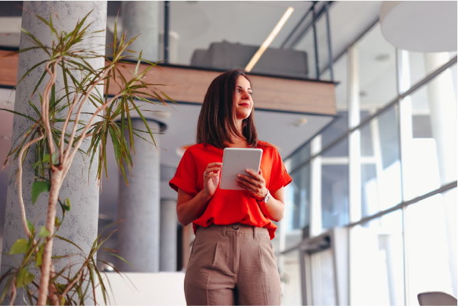Businesswoman is holding a tablet and smiling while looking up in a modern office with a large plant in the foreground